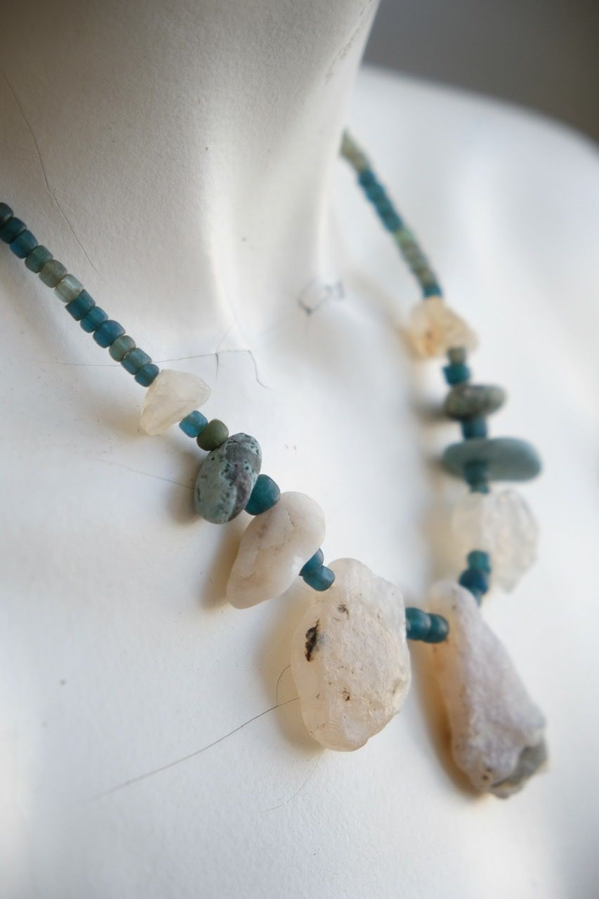 A white colored female form mannequin wears a sculptural necklace of white and blue river tumbled stones and turquoise glass beads