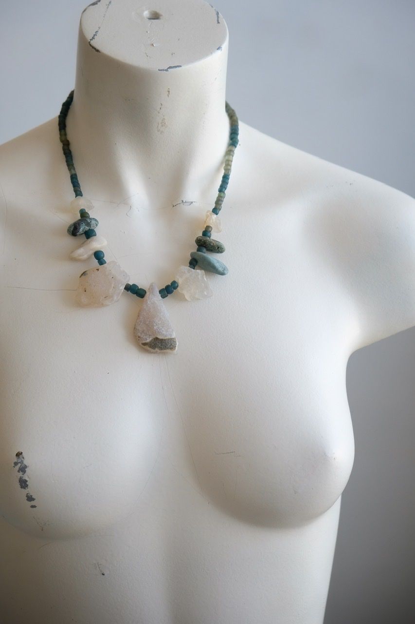 A white colored female form mannequin wears a sculptural necklace of white and blue river tumbled stones and turquoise glass beads