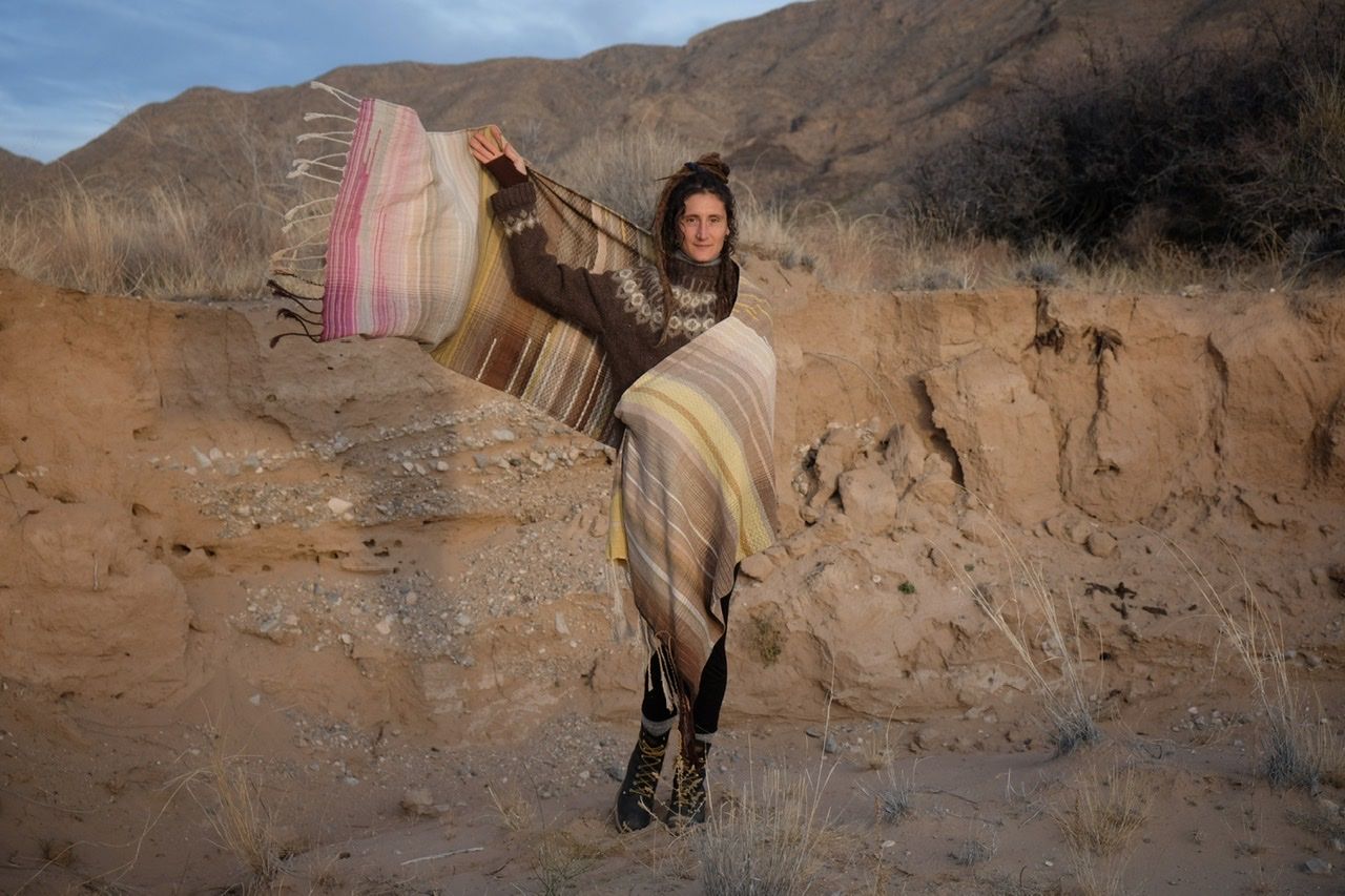 A woman in a brown and tan sweater stands in a sandy desert landscape wearing a shawl