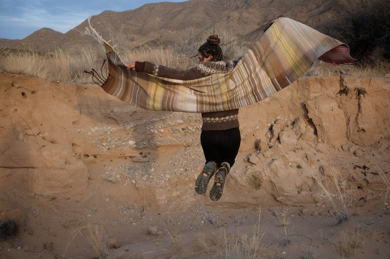 A woman in a brown and tan sweater stands in a sandy desert landscape wearing a shawl