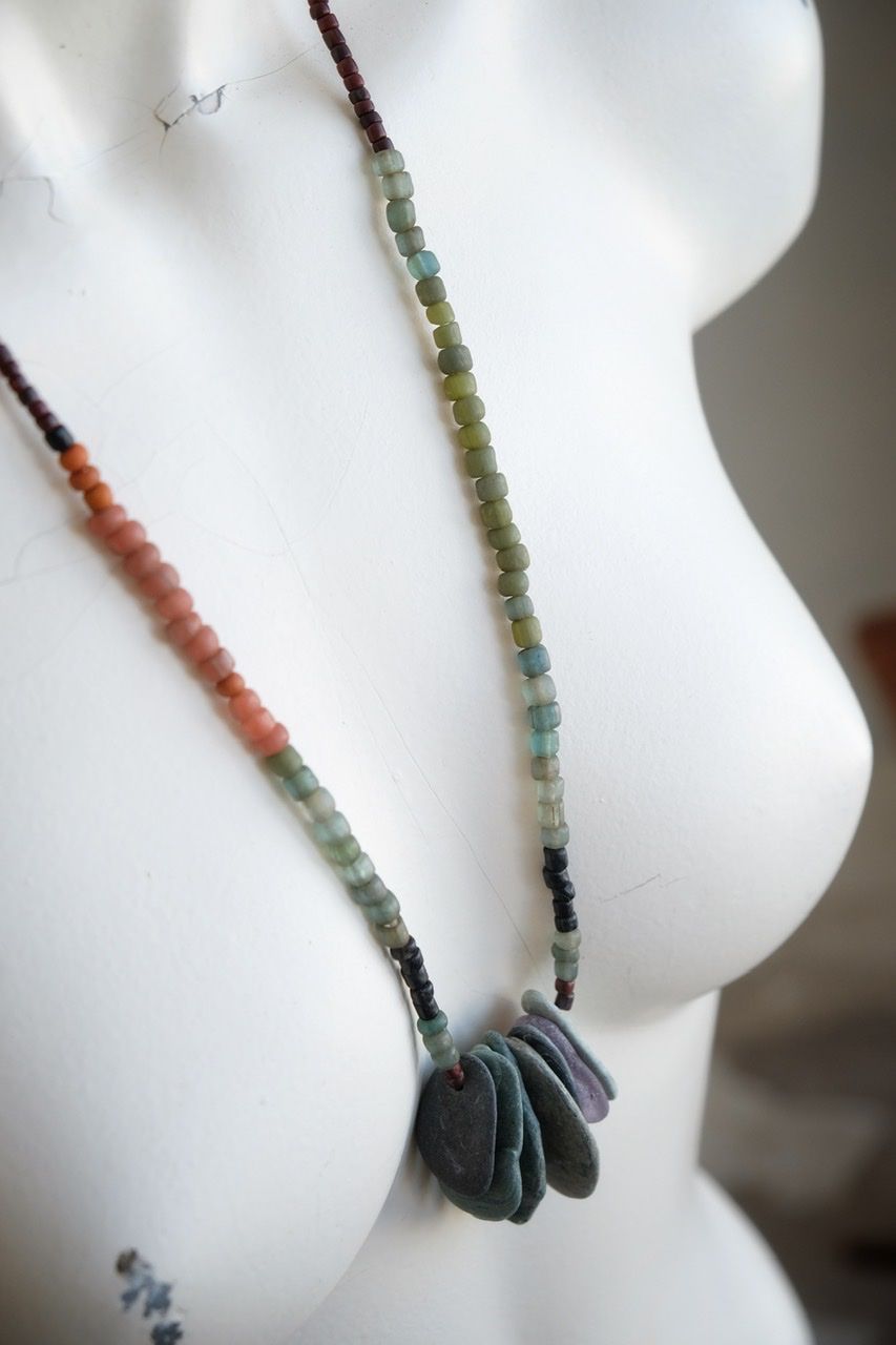 A white colored female form mannequin wears a sculptural necklace of thin grey stones and multicolored glass beads. 