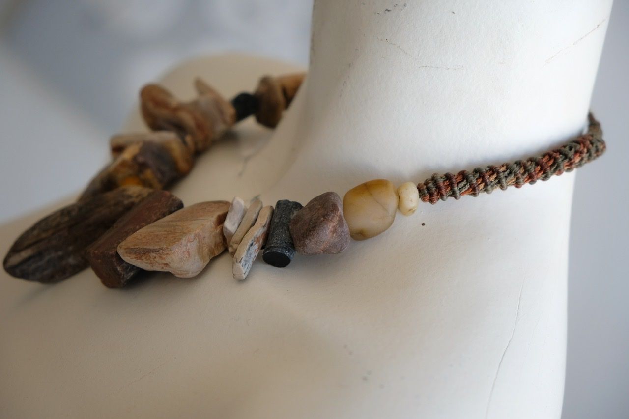A white colored female form mannequin wears a sculptural necklace of petrified wood, smooth stones, yellow glass beads and a knot work tube