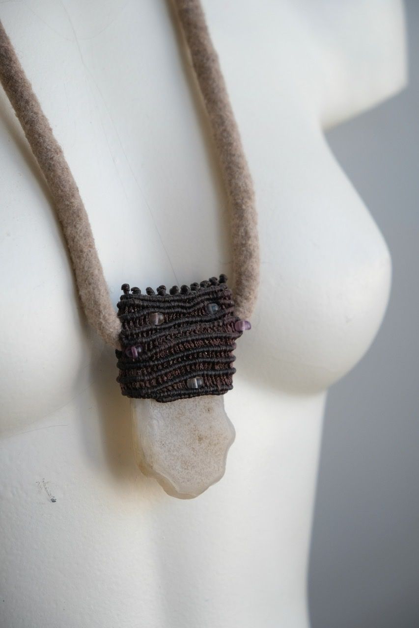 A white colored female form mannequin wears a sculptural necklace of a light grey felted cord, dark purple basket with a white chalcedony stone