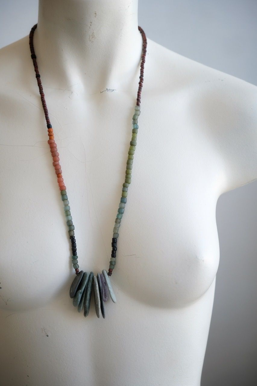 A white colored female form mannequin wears a sculptural necklace of thin grey stones and multicolored glass beads. 