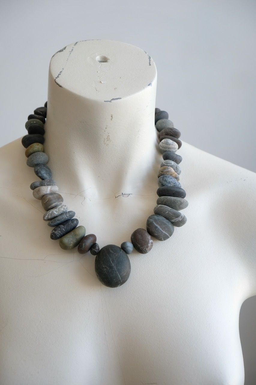 A white color female form mannequin wears a sculptural necklace of smooth dark blue grey stones