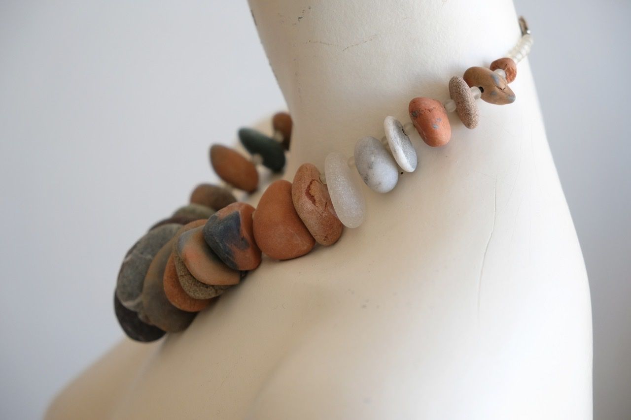 A white female form mannequin wears a sculptural necklace of ocean tumbled stones In greys, brown, red and white