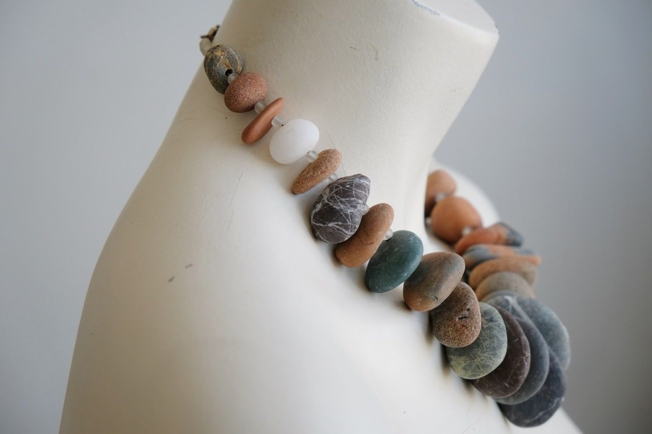 A white female form mannequin wears a sculptural necklace of ocean tumbled stones In greys, brown, red and white