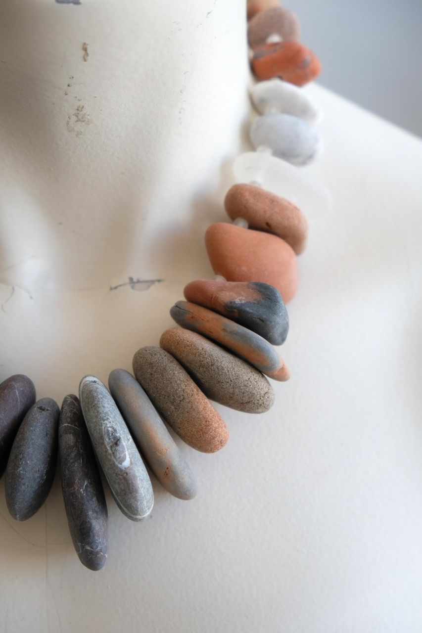 A white female form mannequin wears a sculptural necklace of ocean tumbled stones In greys, brown, red and white