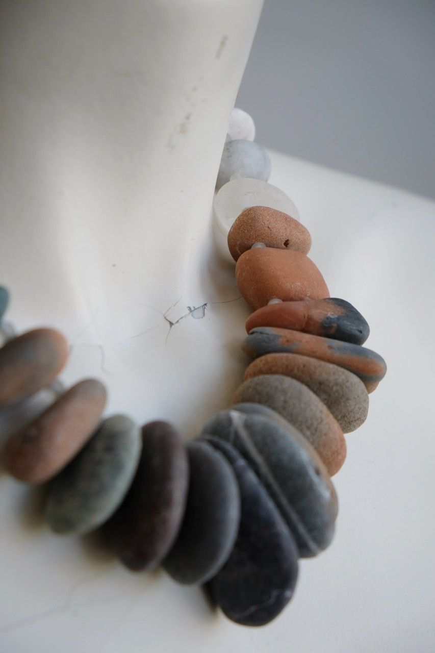 A white female form mannequin wears a sculptural necklace of ocean tumbled stones In greys, brown, red and white