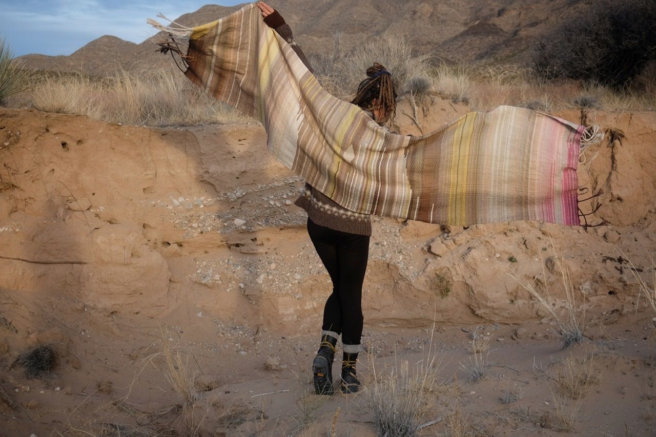 A woman in a brown and tan sweater stands in a sandy desert landscape wearing a shawl