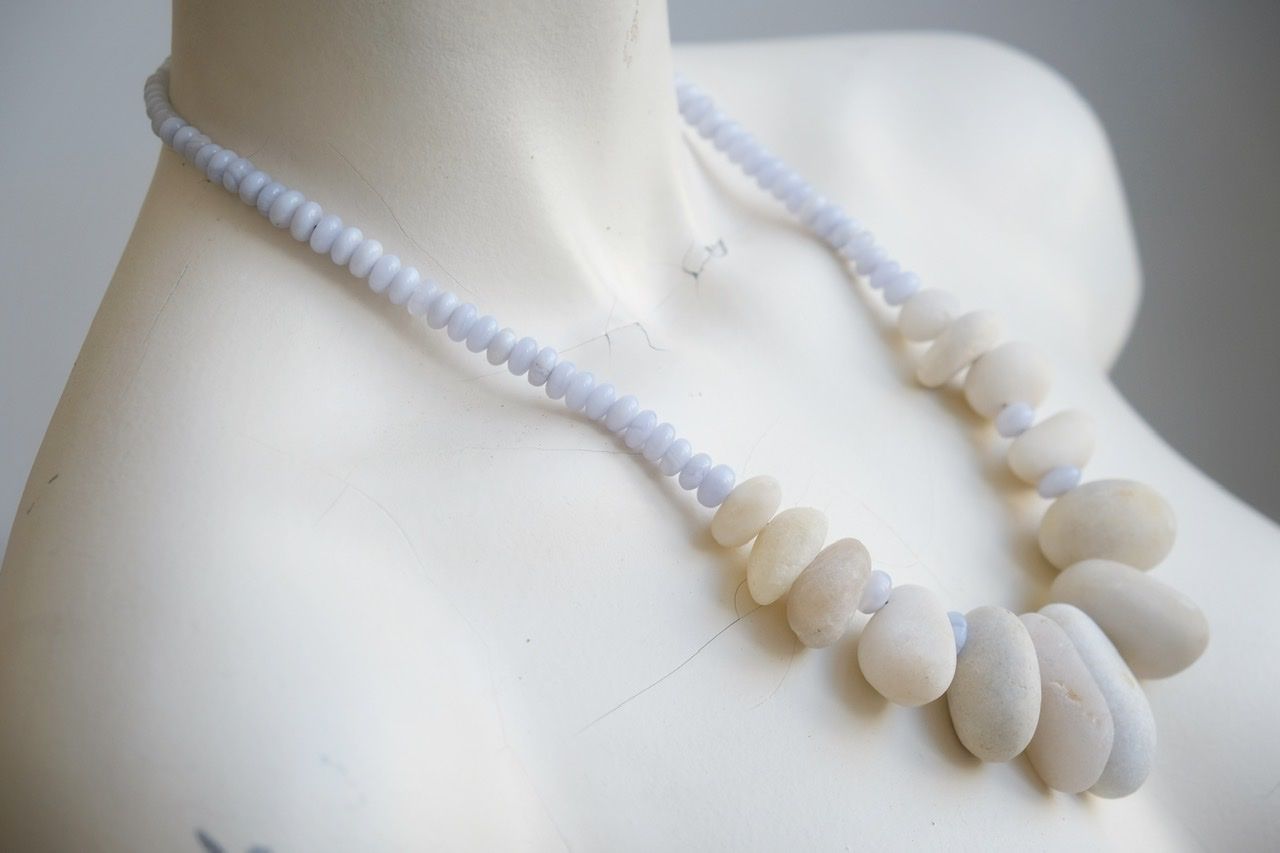 A white colored female form mannequin wears a sculptural necklace of white stone orbs and crystalline blue chalcedony beads