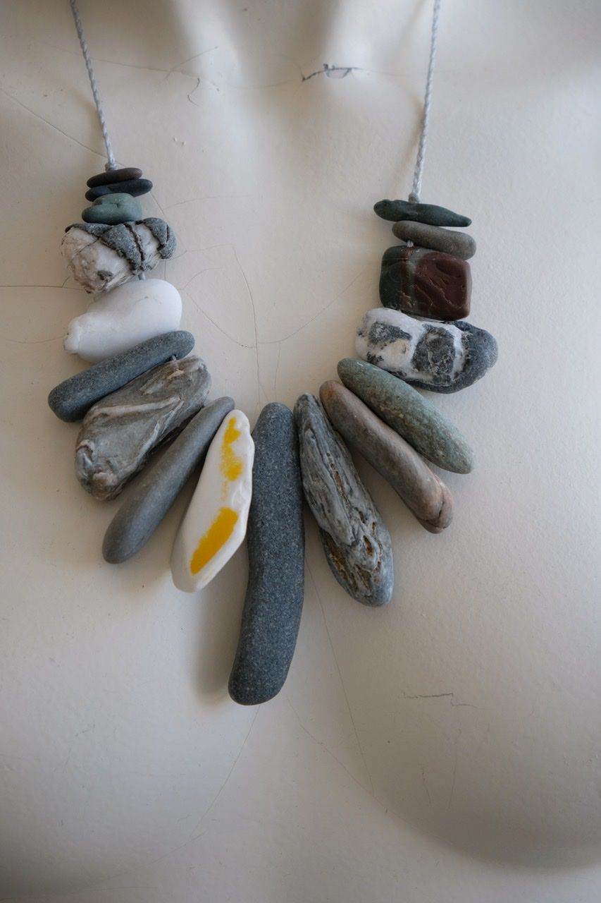 A white colored female form mannequin wears a sculptural necklace of gray braided twine with rough stones hanging from it.