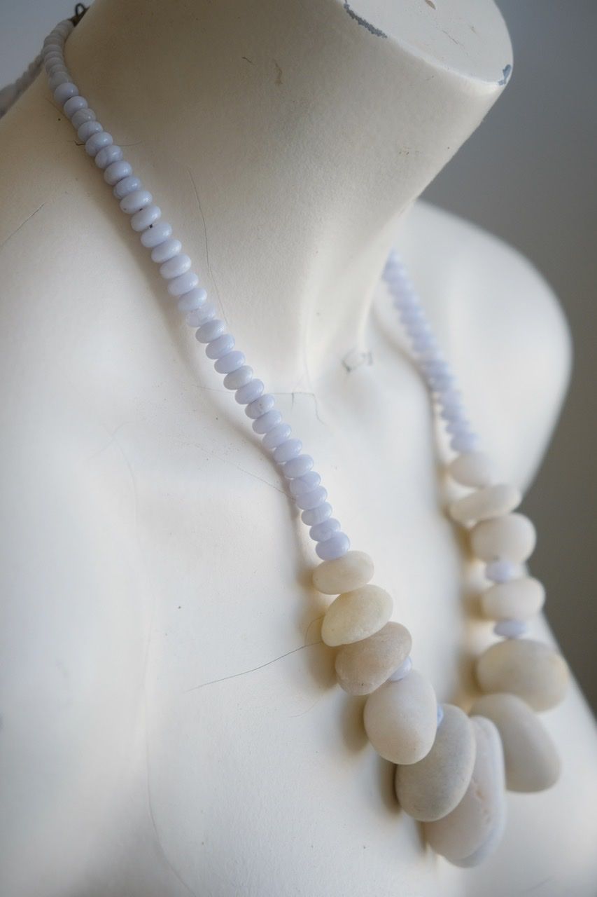 A white colored female form mannequin wears a sculptural necklace of white stone orbs and crystalline blue chalcedony beads