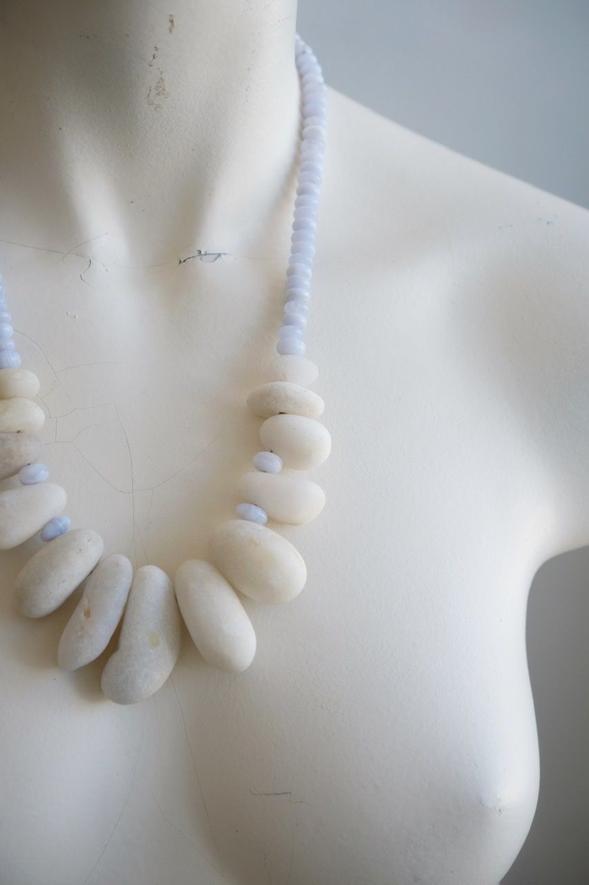 A white colored female form mannequin wears a sculptural necklace of white stone orbs and crystalline blue chalcedony beads