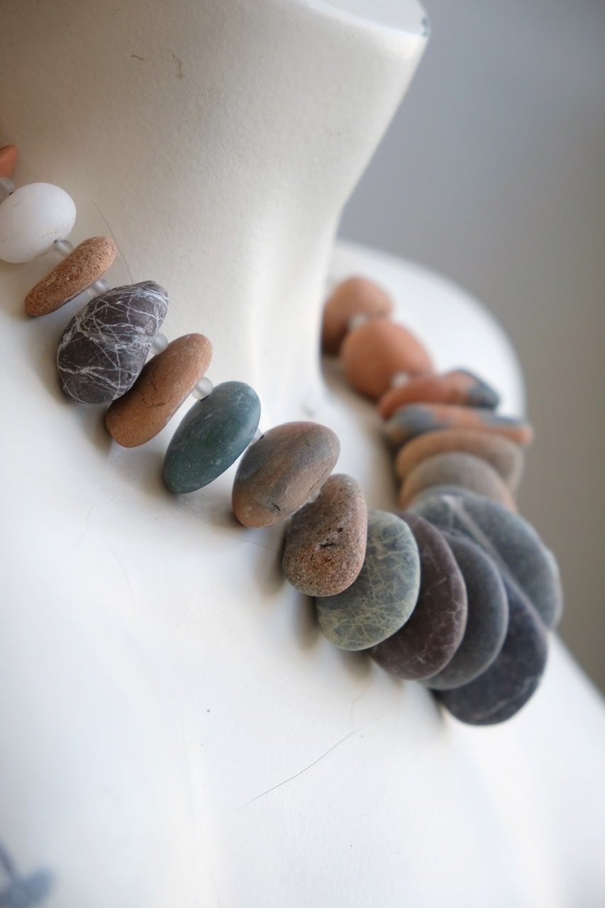 A white female form mannequin wears a sculptural necklace of ocean tumbled stones In greys, brown, red and white