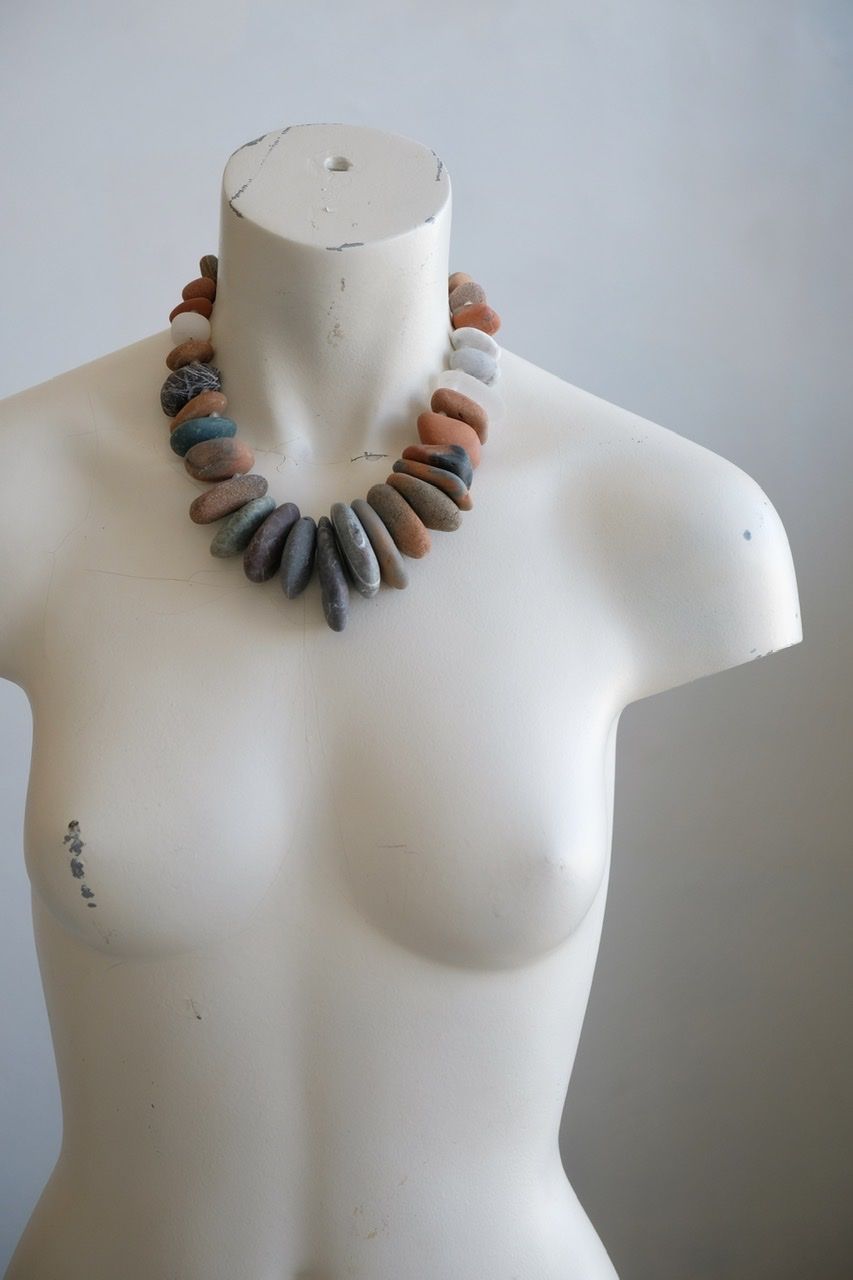 A white female form mannequin wears a sculptural necklace of ocean tumbled stones In greys, brown, red and white