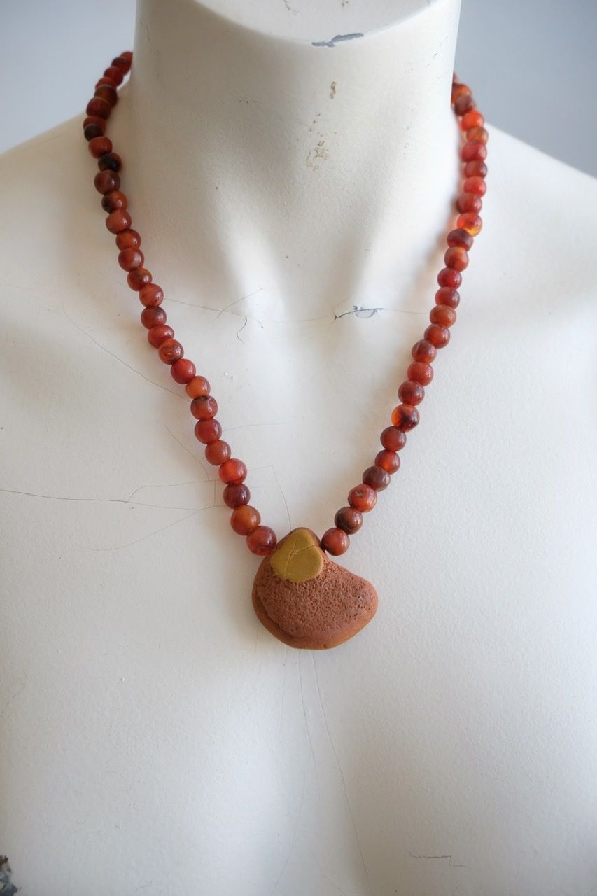 A white color female form mannequin wears a sculptural stone necklace in red, orange and yellow ochre colors 