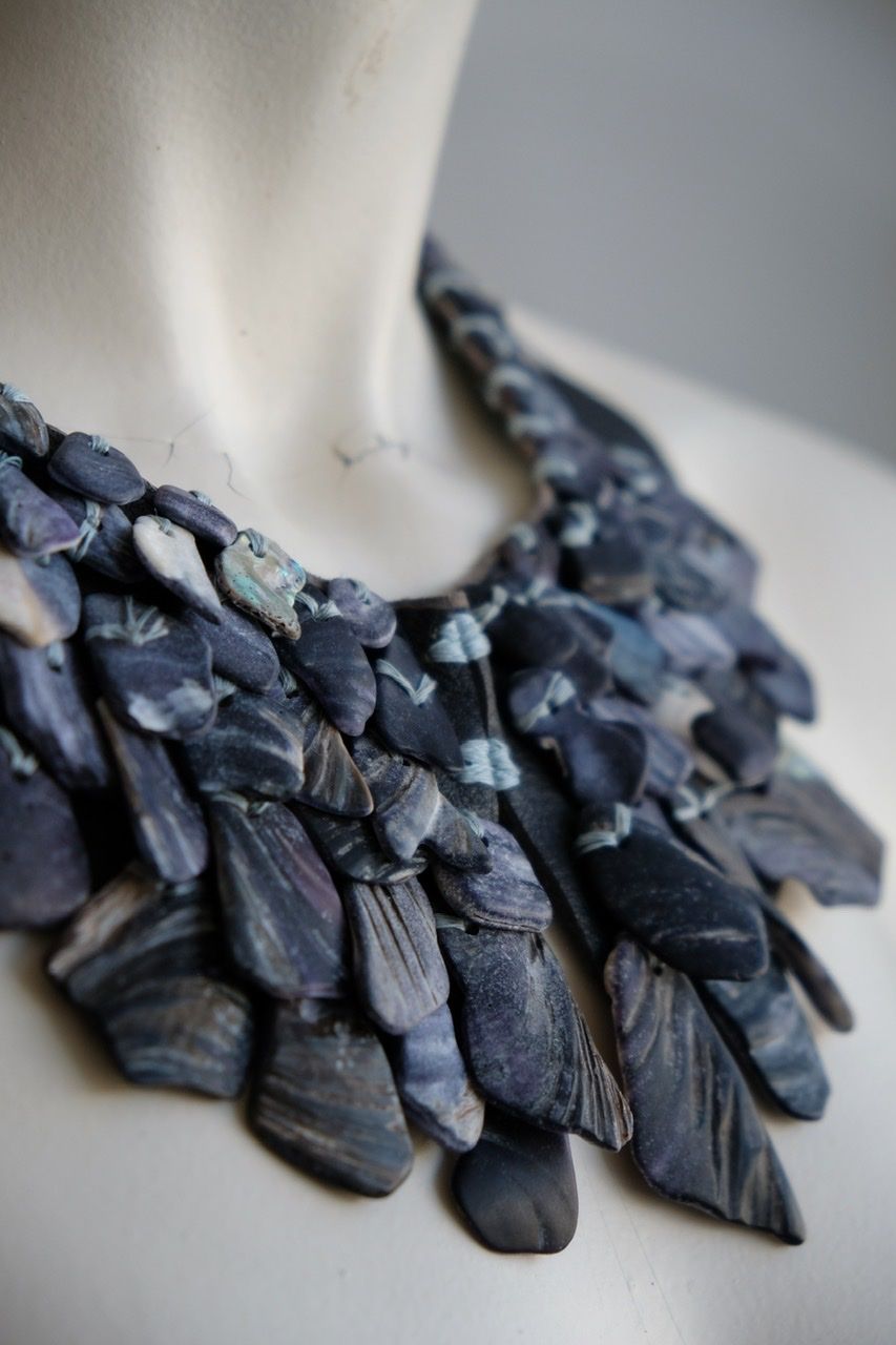 A white female formed mannequin wears a sculptural necklace of blue shells and blue leather