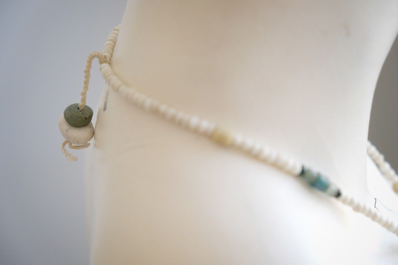 A white colored female form mannequin wears a sculptural necklace of blue green river tumbled stones and white glass beads