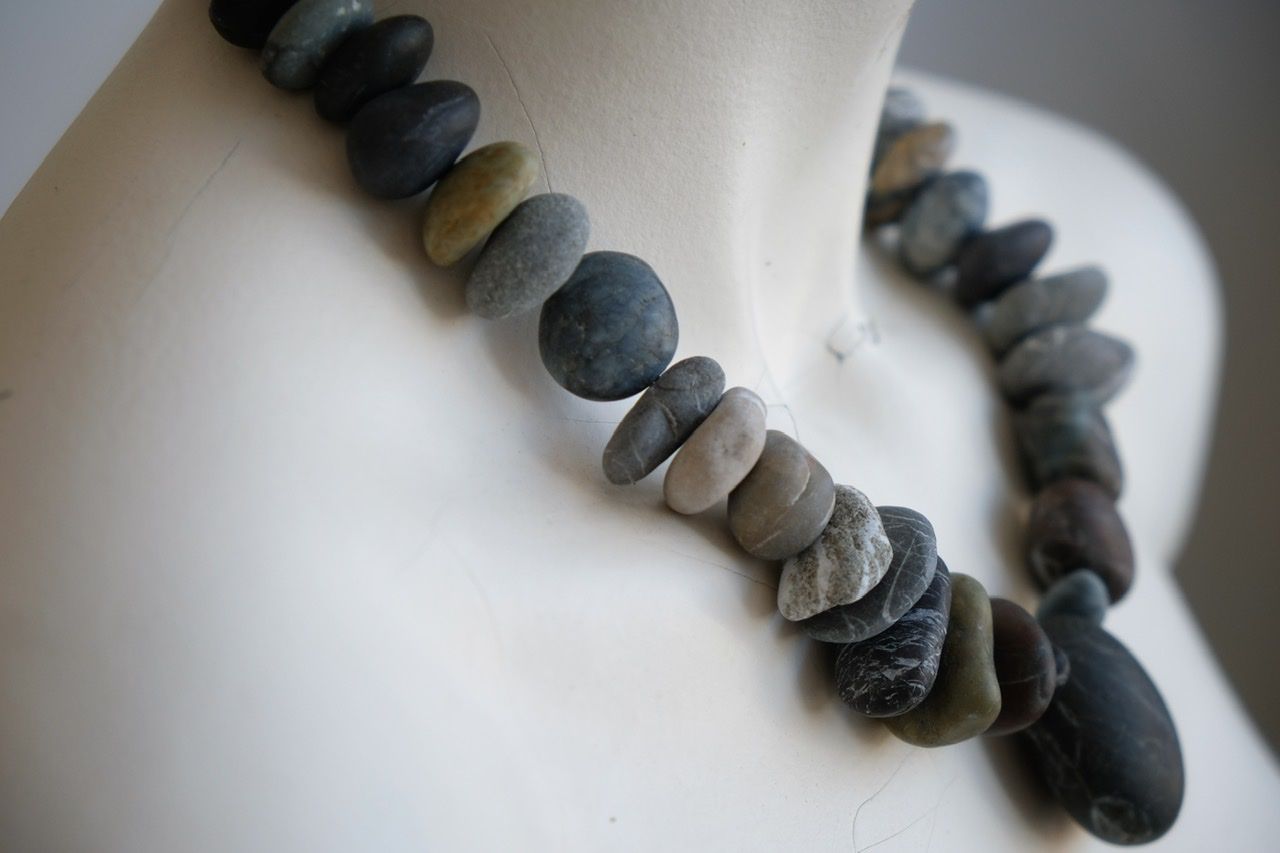 A white color female form mannequin wears a sculptural necklace of smooth dark blue grey stones