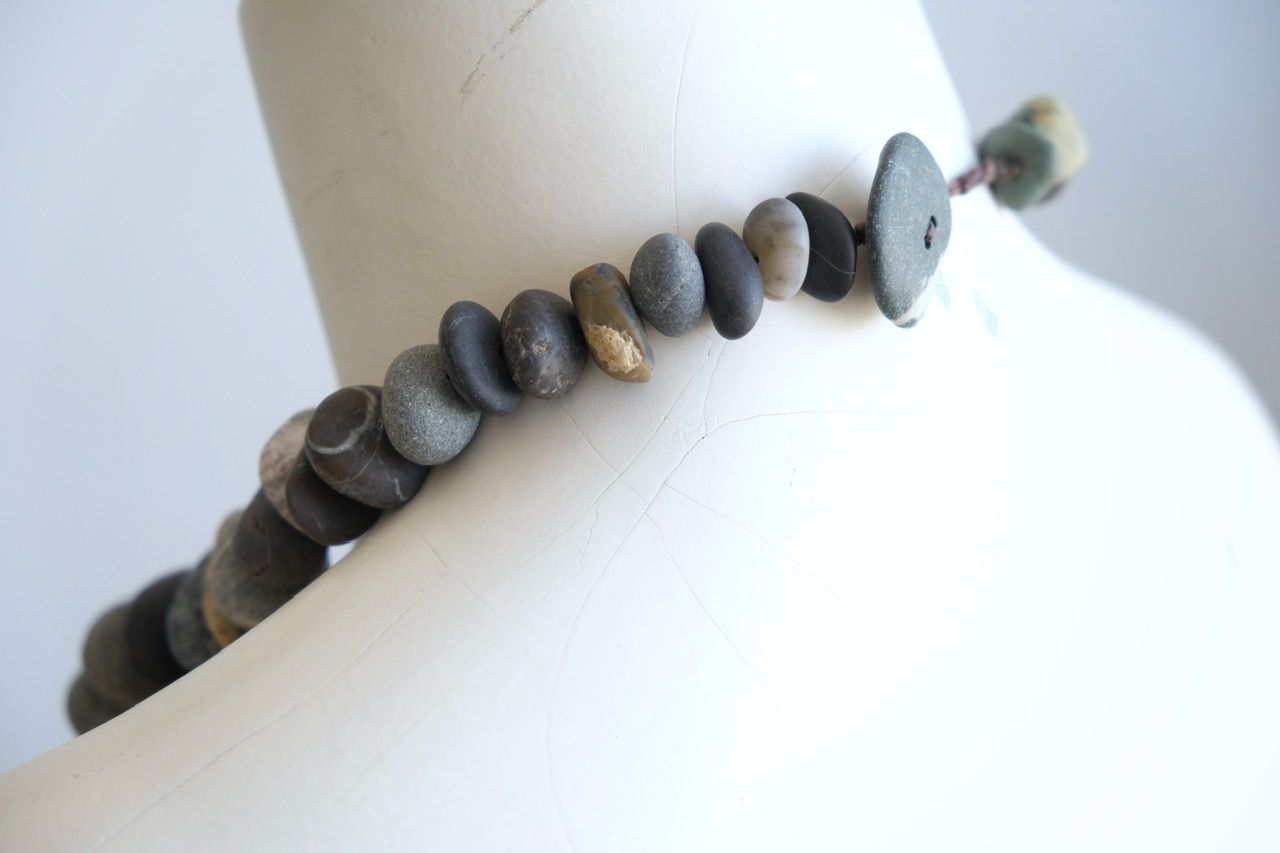 A white color female form mannequin wears a sculptural necklace of smooth dark blue grey stones