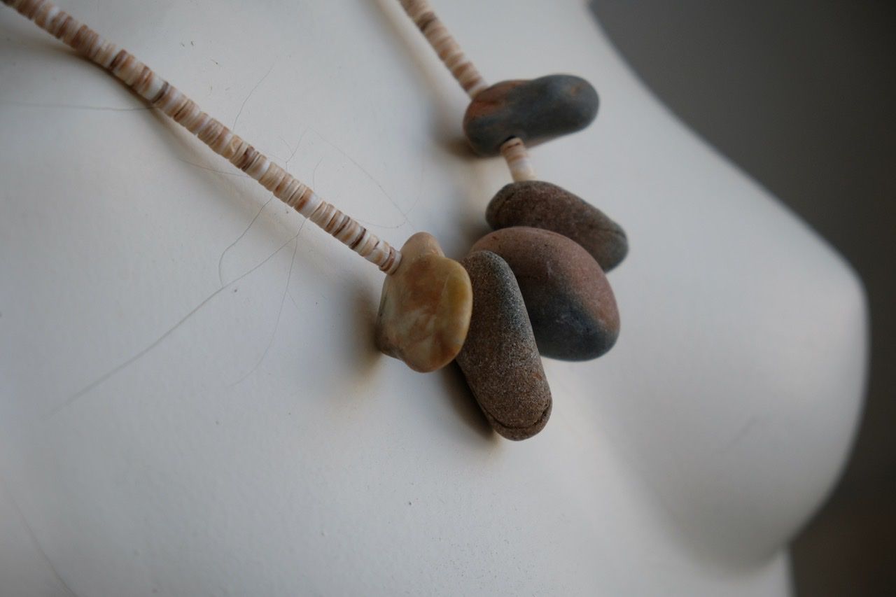 A white female form mannequin wears a sculptural necklace of ocean tumbled stones In greys, brown, red and white