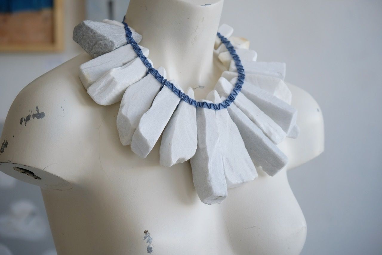 A white colored female form mannequin wears a sculptural necklace of large wedges of white marble and blue string
