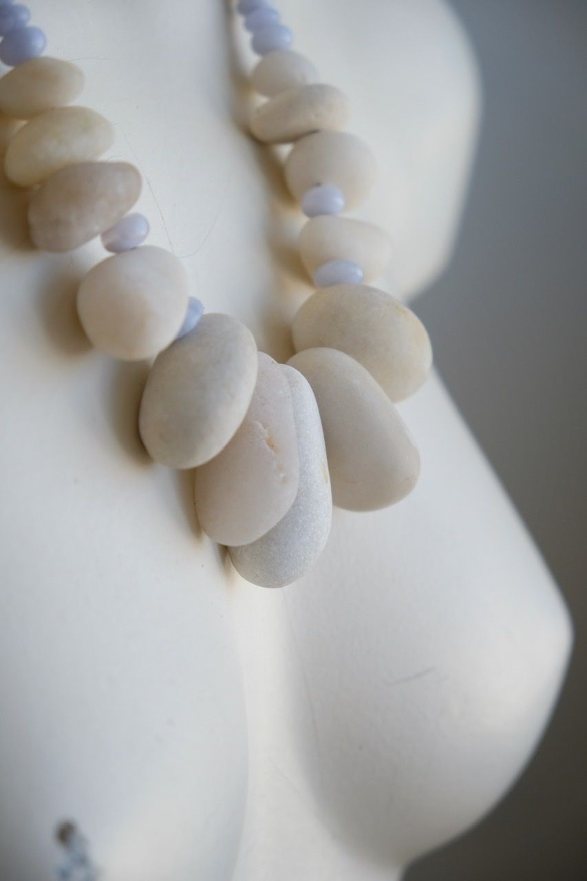 A white colored female form mannequin wears a sculptural necklace of white stone orbs and crystalline blue chalcedony beads