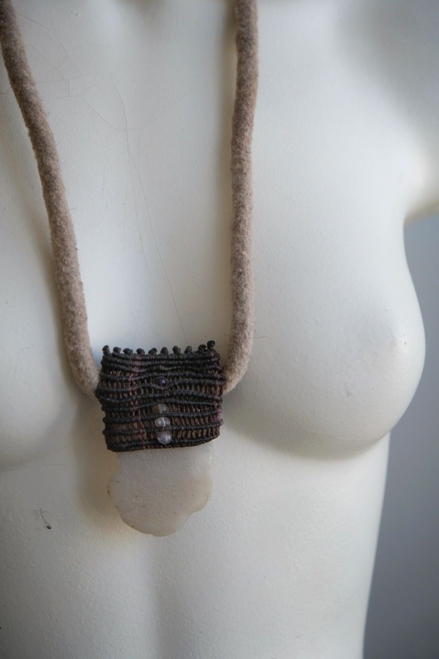 A white colored female form mannequin wears a sculptural necklace of a light grey felted cord, dark purple basket with a white chalcedony stone