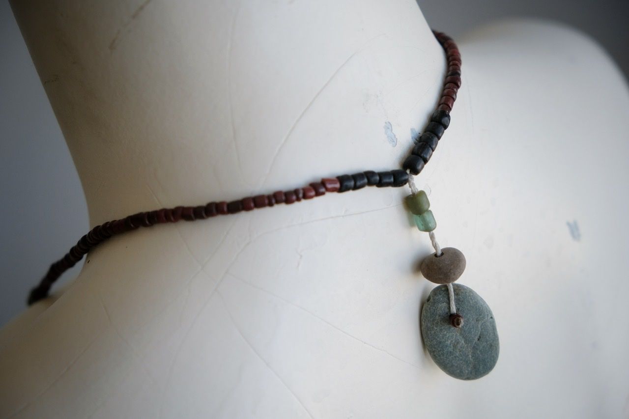 A white colored female form mannequin wears a sculptural necklace of thin grey stones and multicolored glass beads. 