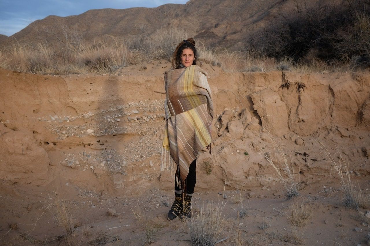 A woman in a brown and tan sweater stands in a sandy desert landscape wearing a shawl
