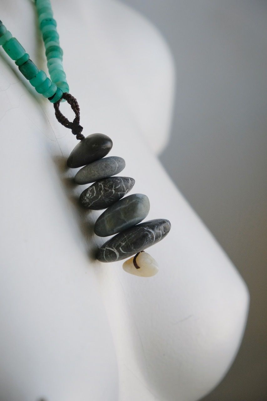 A white color female form mannequin wears a sculptural necklace of smooth dark blue grey stones In the form of a tiny cairn