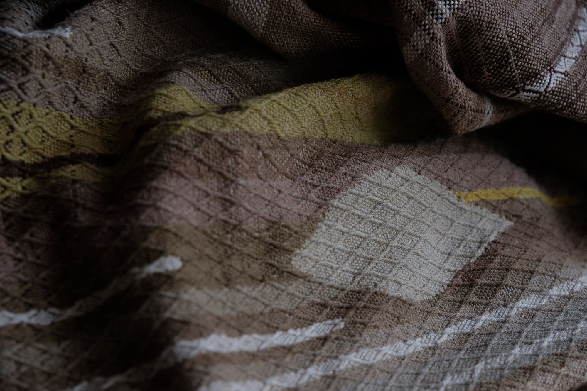 detail of handwoven diamond pattern and geometric details in browns, tan, yellows, whites and pinks laying on a wooden floor