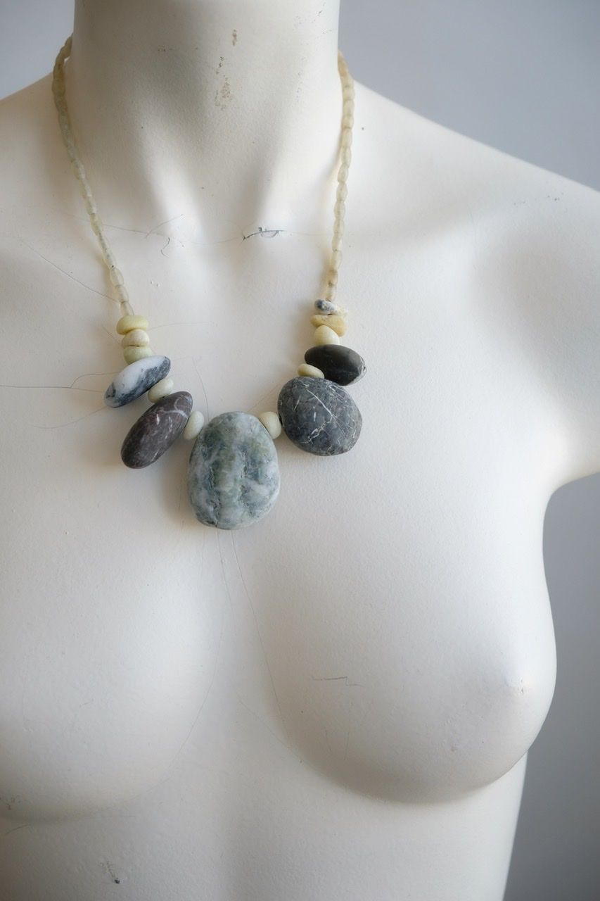 A white color female form mannequin wears a sculptural necklace of smooth dark blue grey stones