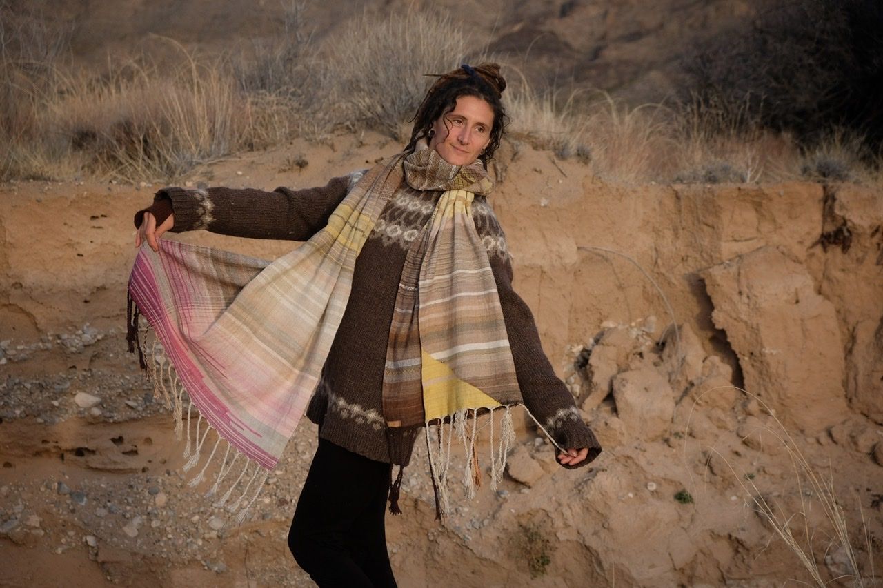 A woman in a brown and tan sweater stands in a sandy desert landscape wearing a shawl