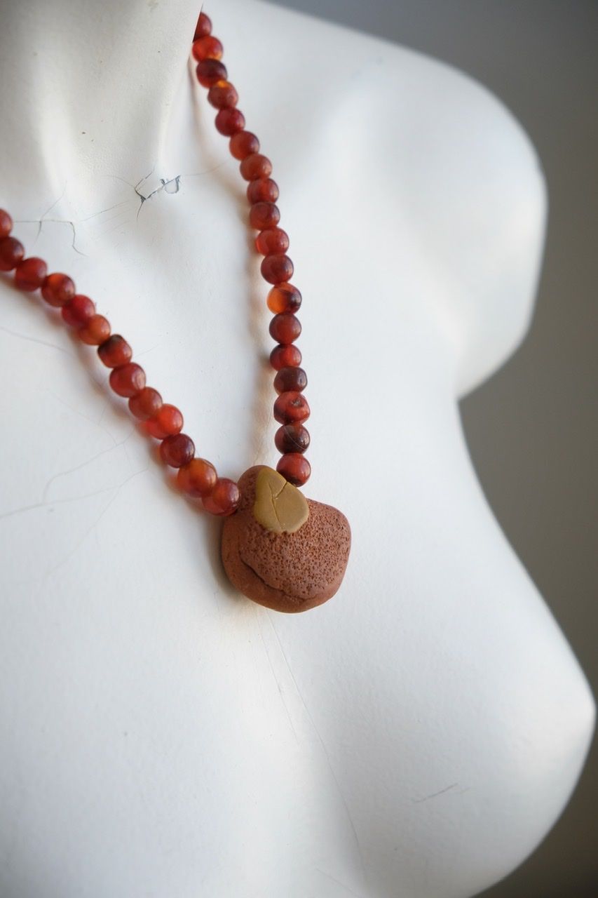 A white color female form mannequin wears a sculptural stone necklace in red, orange and yellow ochre colors 