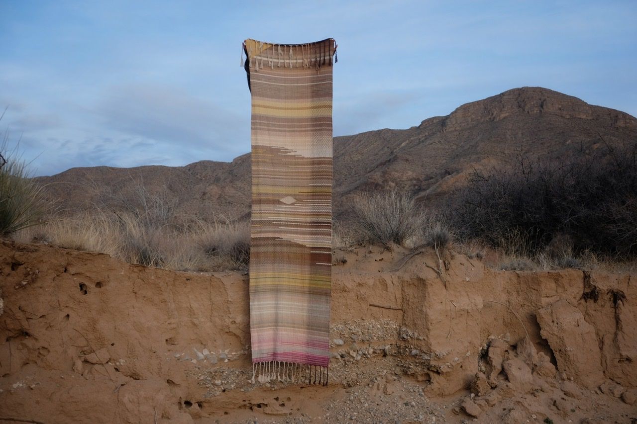 A woman in a brown and tan sweater stands in a sandy desert landscape wearing a shawl