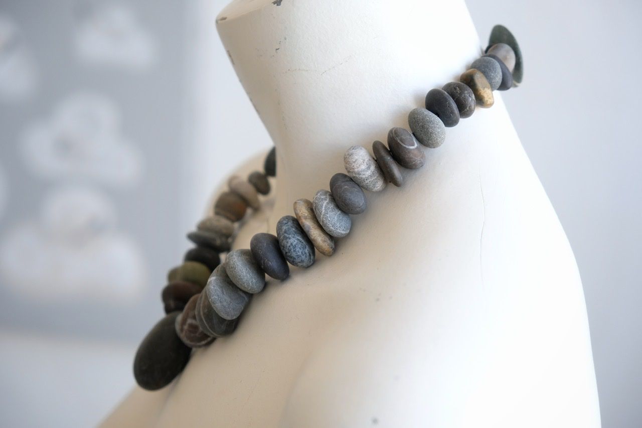 A white color female form mannequin wears a sculptural necklace of smooth dark blue grey stones