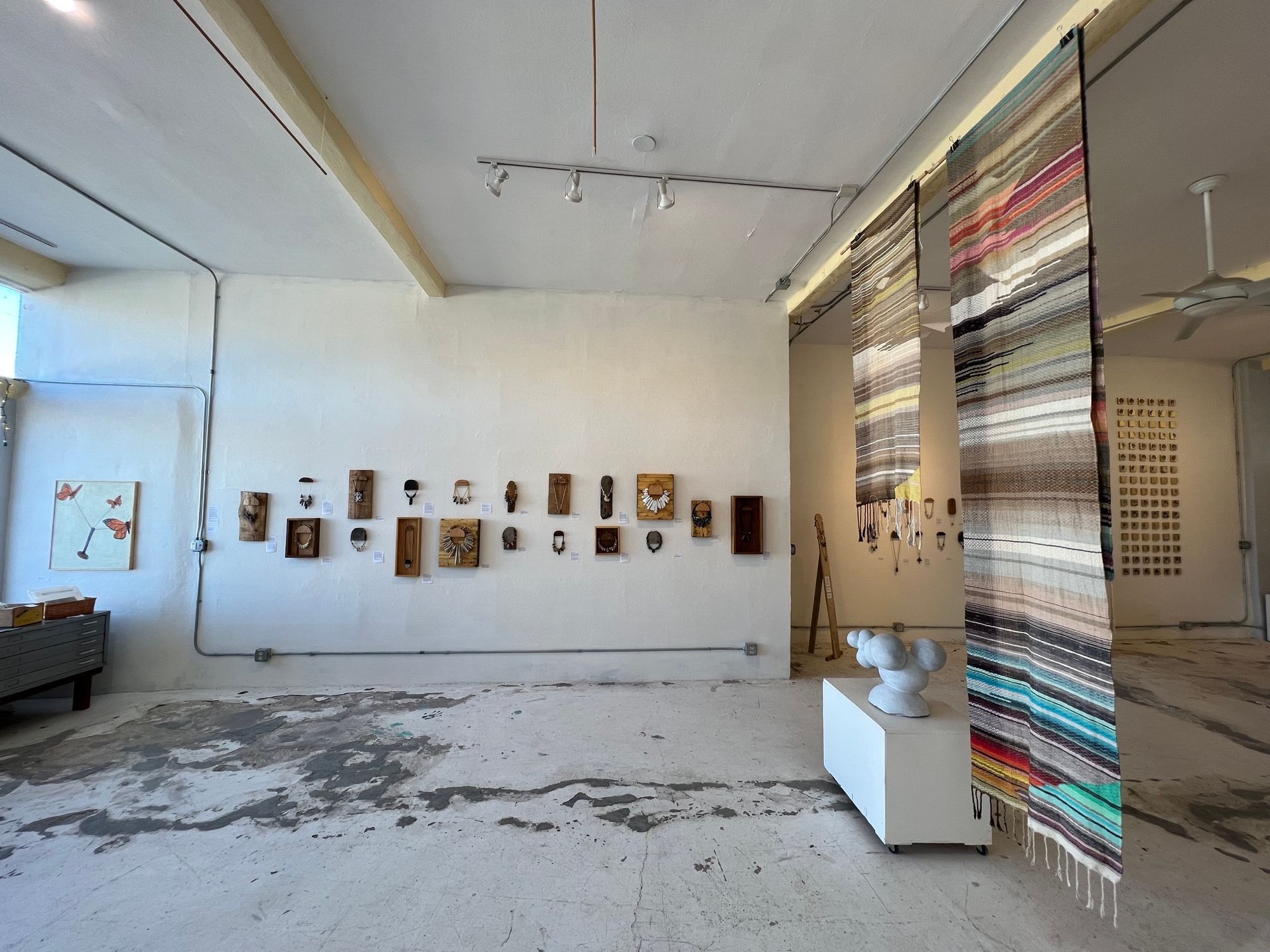 Handwoven natural and rainbow colored fabric hanging in the middle of a gallery with art on the white walls all around