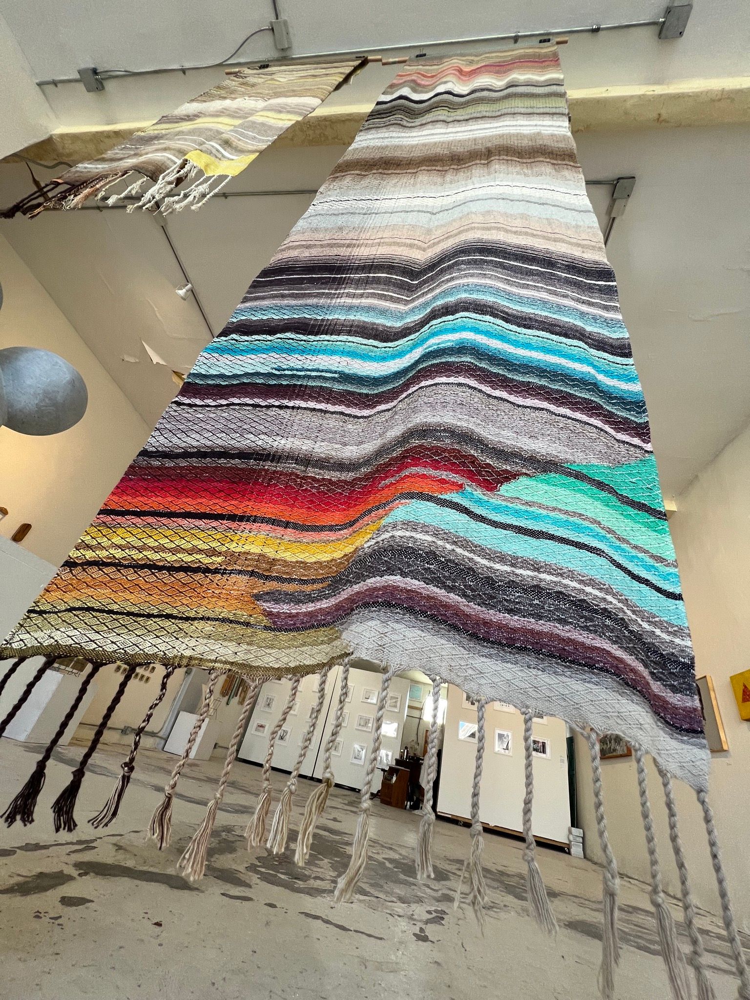 Handwoven natural and rainbow colored fabric hanging in the middle of a gallery with art on the white walls all around