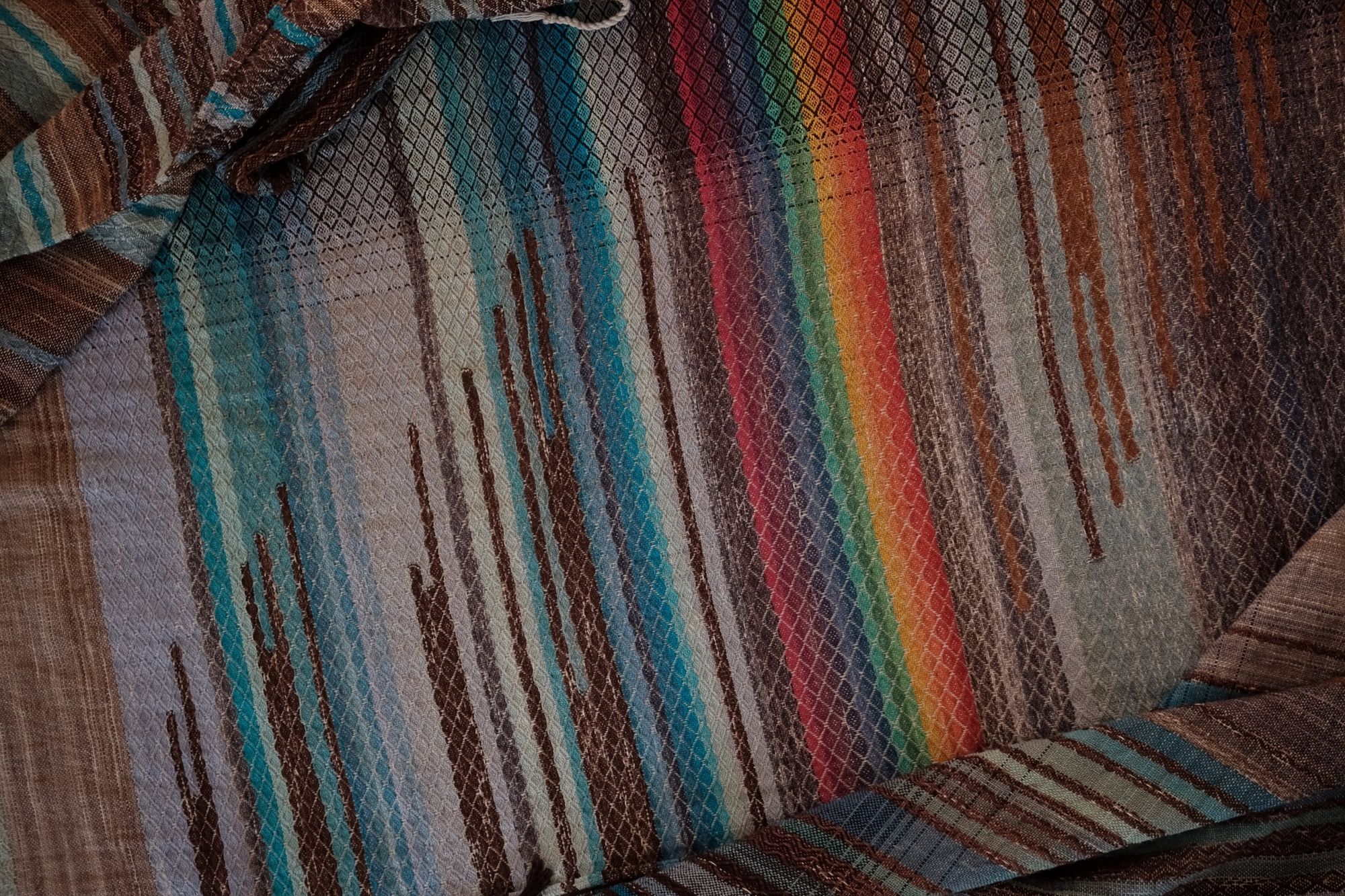 Handwoven fabric with a diamond weave pattern in shades of brown, blue and rainbow lays on a wooden floor