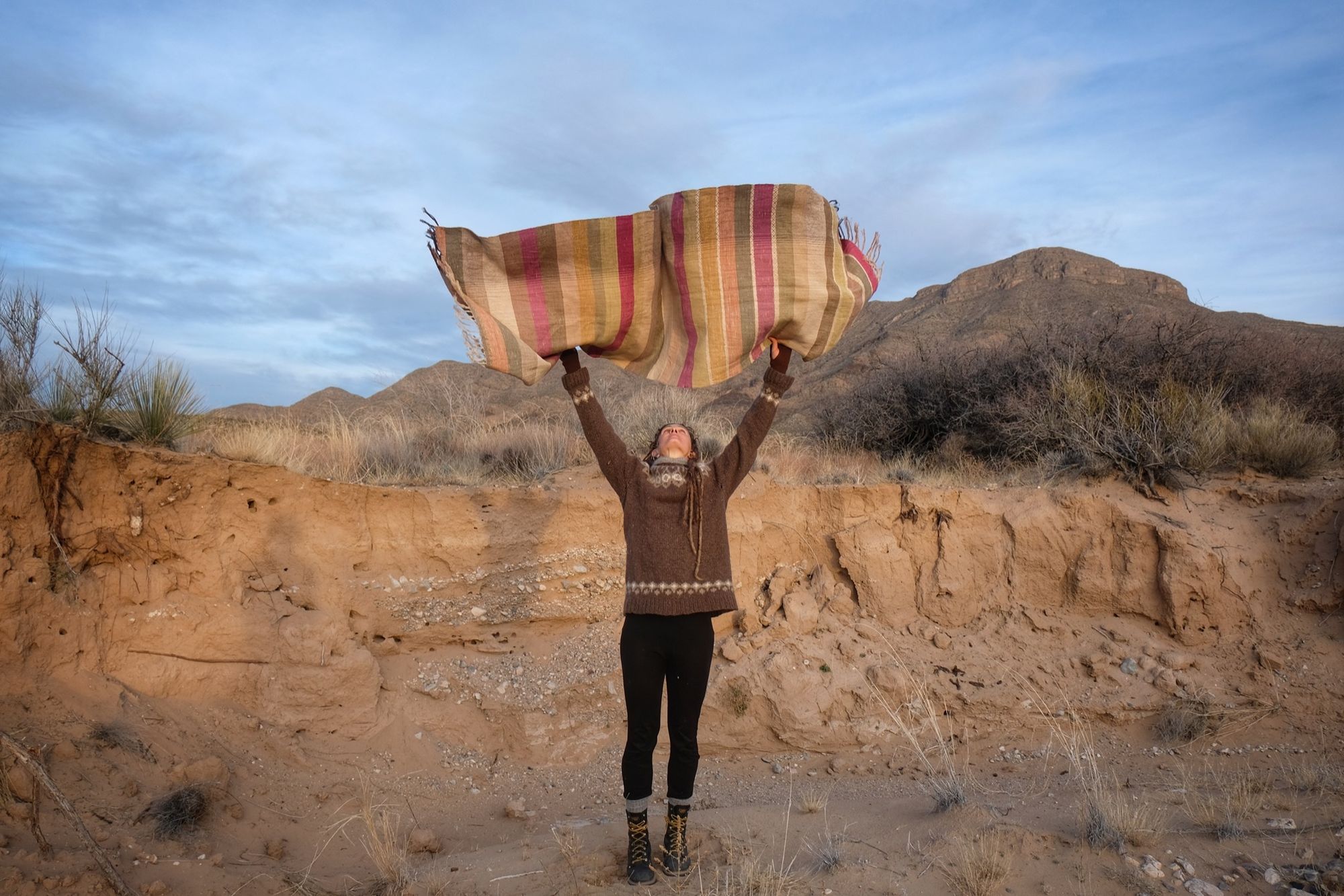 A woman wearing a handwoven silk fabric in soft rainbow striped shades, naturally dyed, in a desert landscape