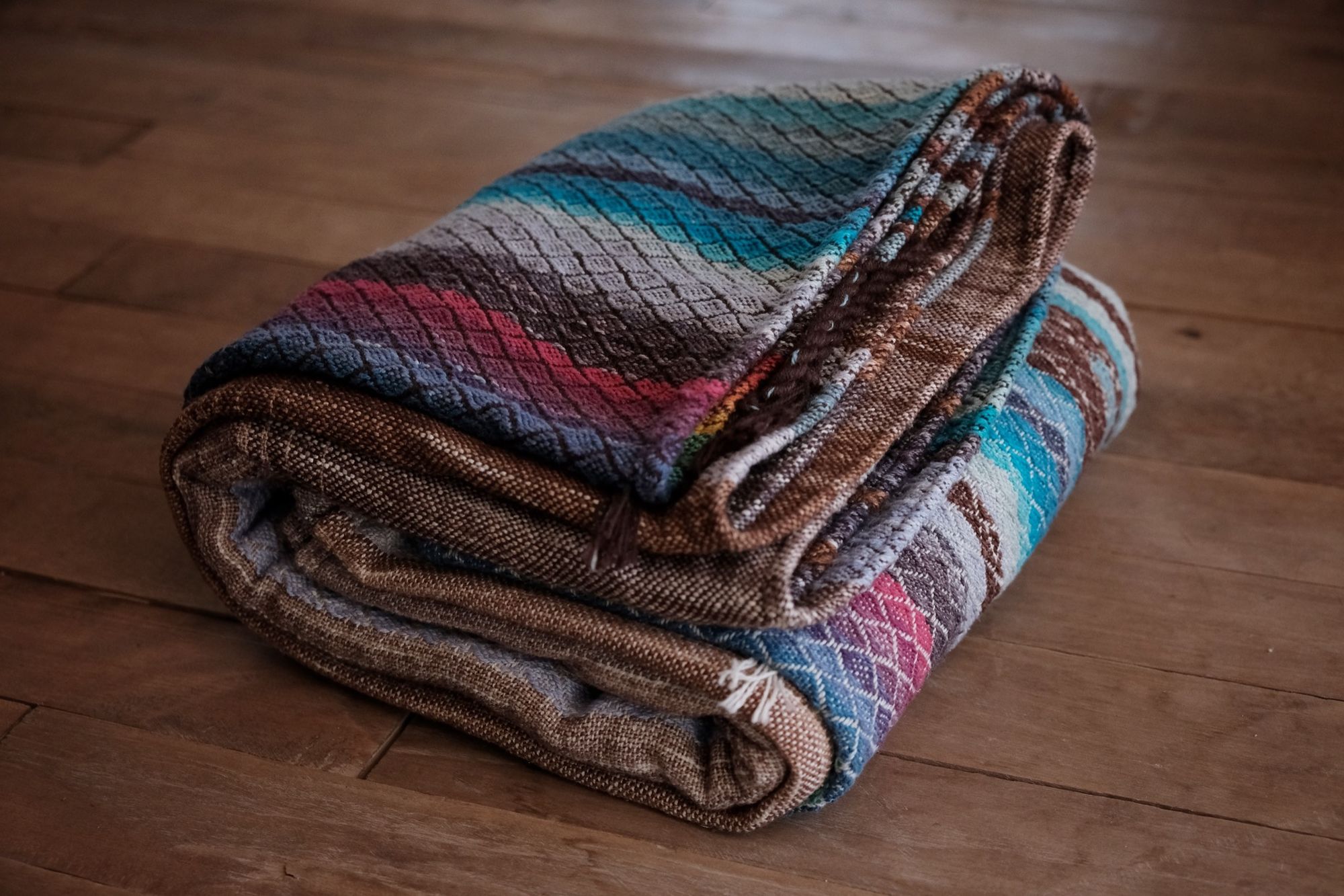 Handwoven fabric with a diamond weave pattern in shades of brown, blue and rainbow lays on a wooden floor