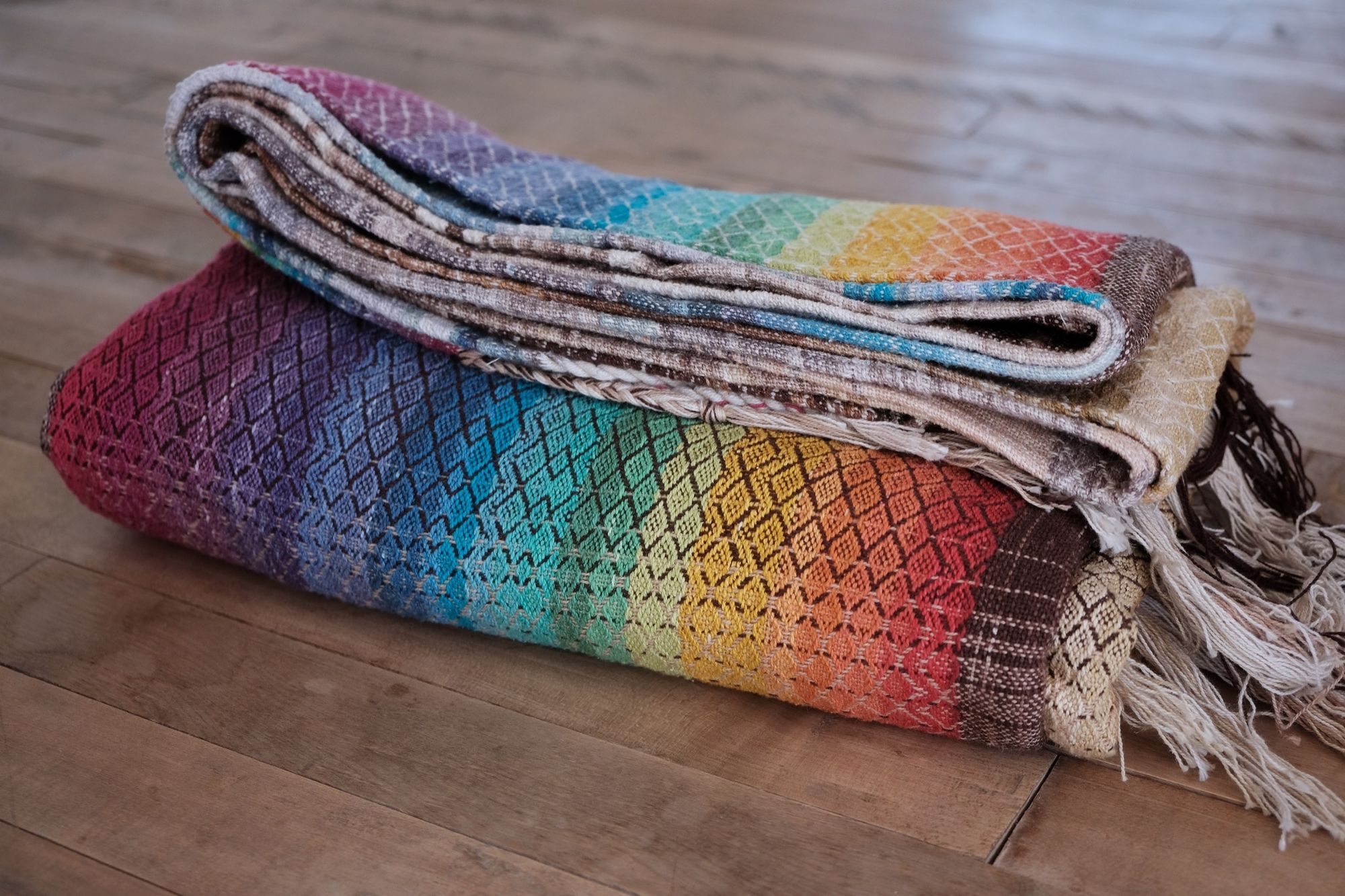 Handwoven fabric with a diamond pattern in brown, cream, white and rainbow colors folded on a wood floor
