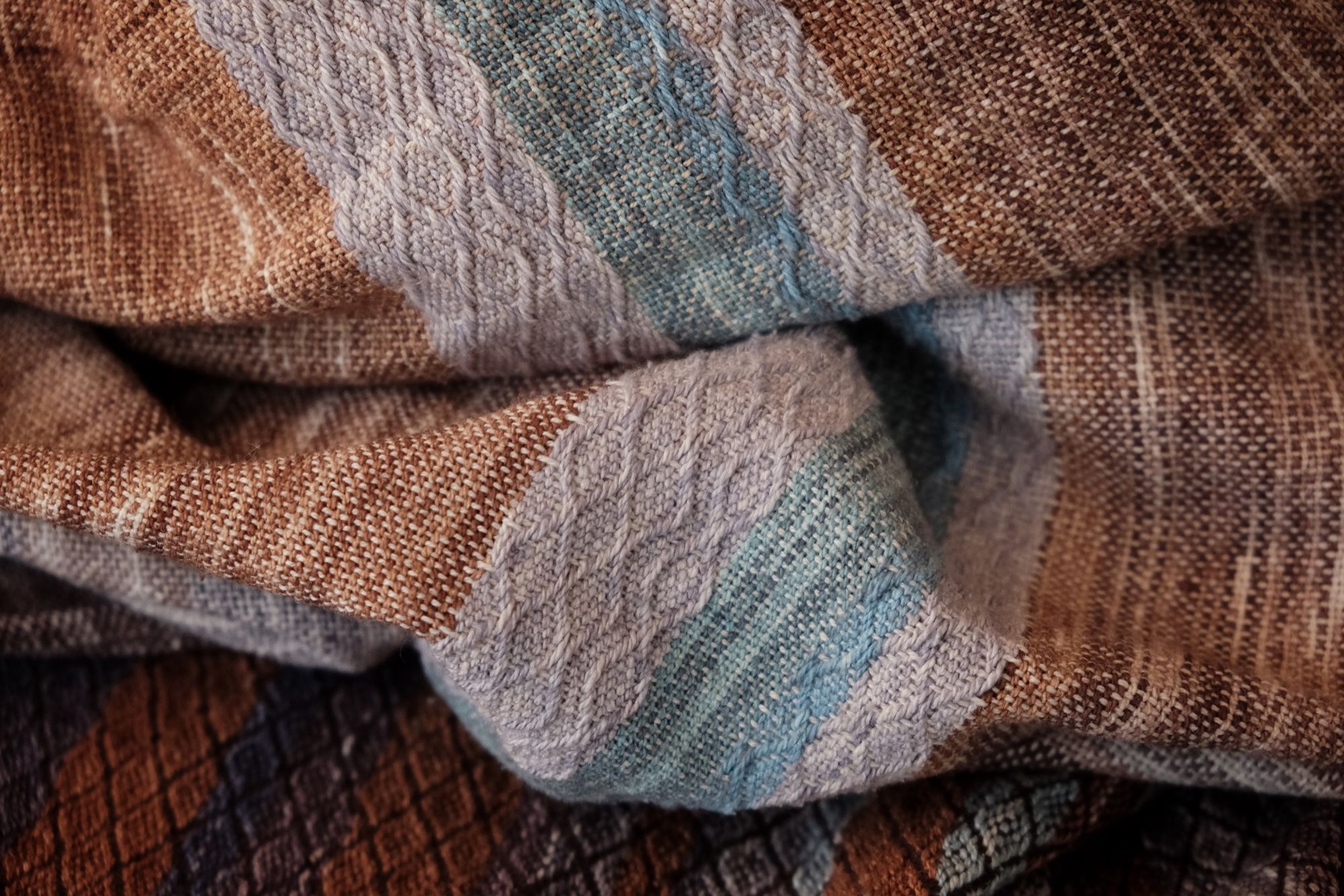 Handwoven fabric with a diamond weave pattern in shades of brown, blue and rainbow lays on a wooden floor