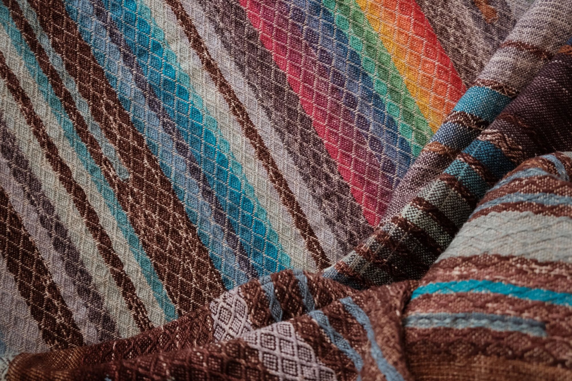 Handwoven fabric with a diamond weave pattern in shades of brown, blue and rainbow lays on a wooden floor