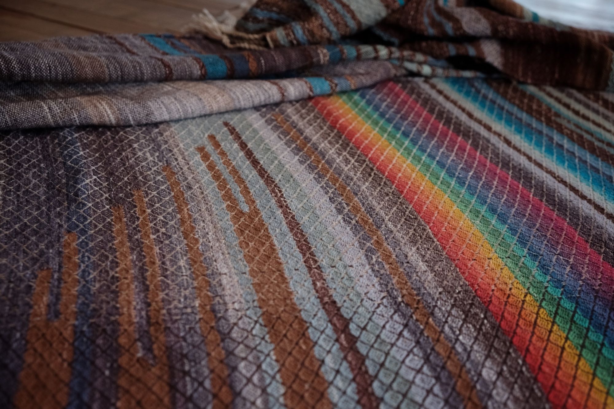 Handwoven fabric with a diamond weave pattern in shades of brown, blue and rainbow lays on a wooden floor