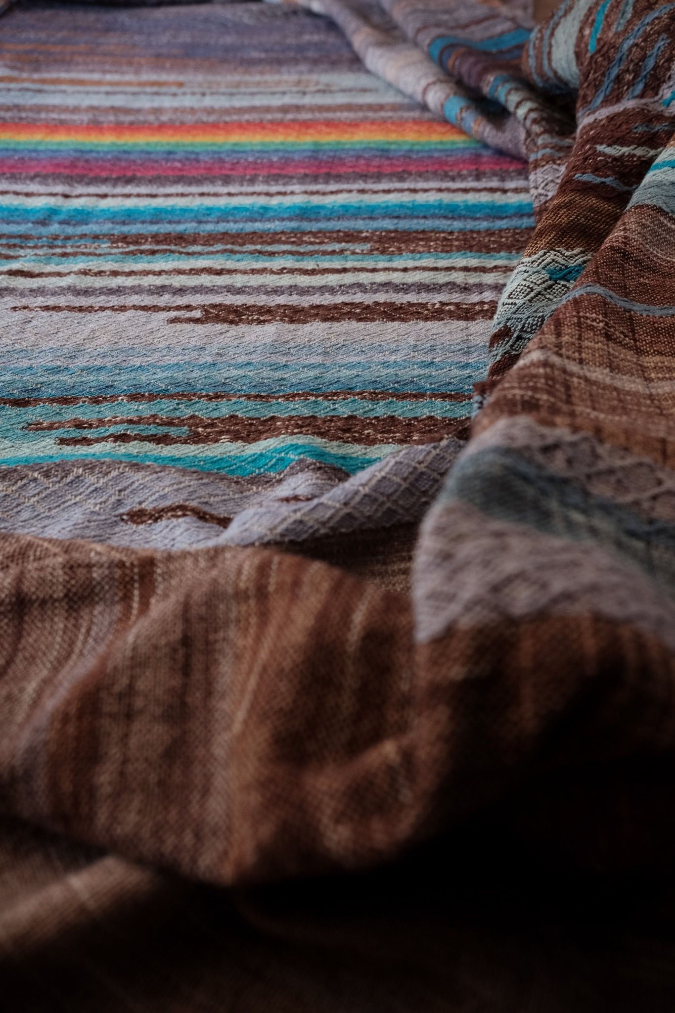 Handwoven fabric with a diamond weave pattern in shades of brown, blue and rainbow lays on a wooden floor