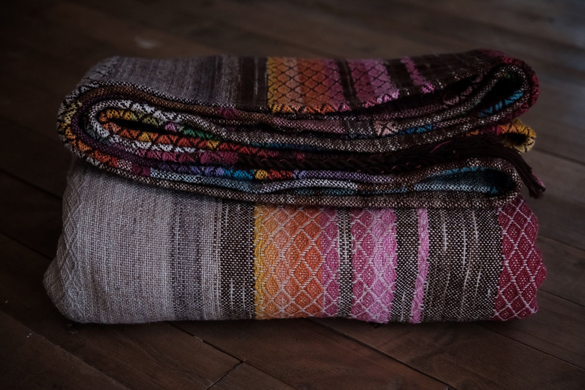 handwoven raw silk fabric woven with a diamond pattern in rainbow, gray, brown and cream colors folded on a wooden floor