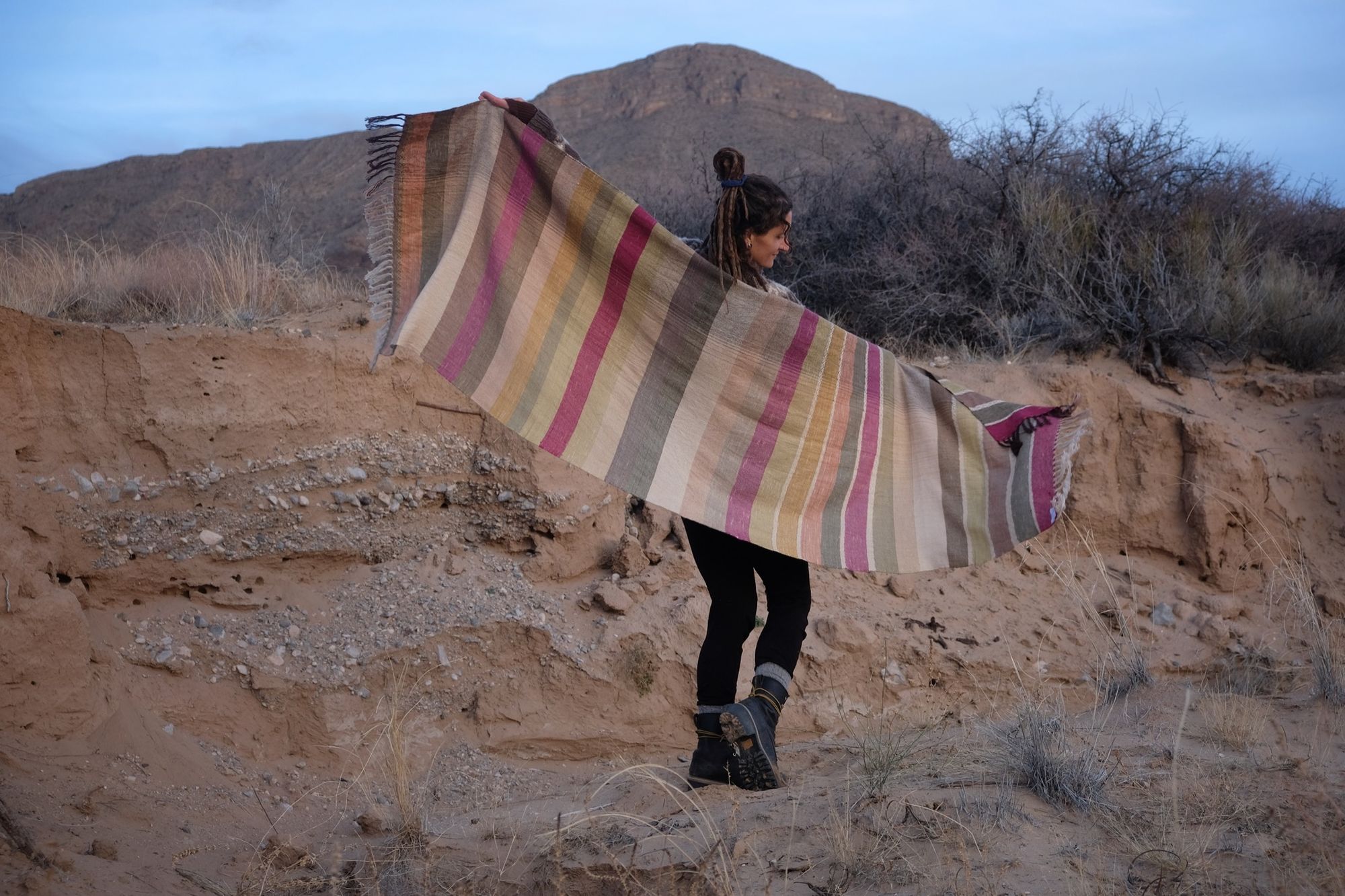 A woman wearing a handwoven silk fabric in soft rainbow striped shades, naturally dyed, in a desert landscape